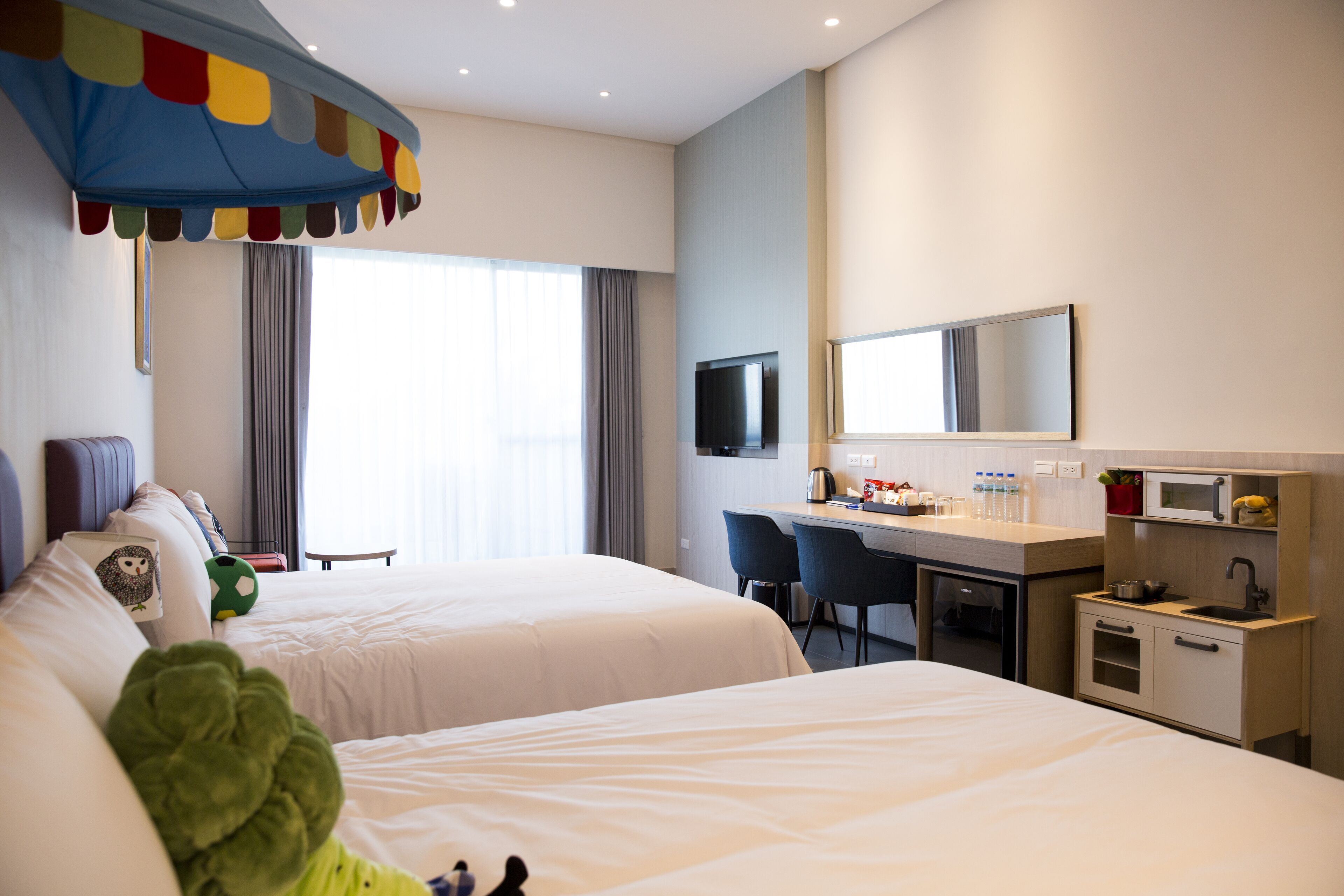 A hotel room with two beds, a desk, a television, and a kitchenette.