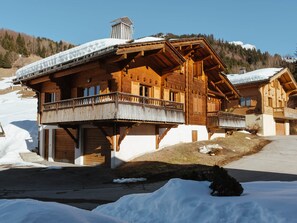 A wooden chalet with a balcony, set against a snowy mountain backdrop.