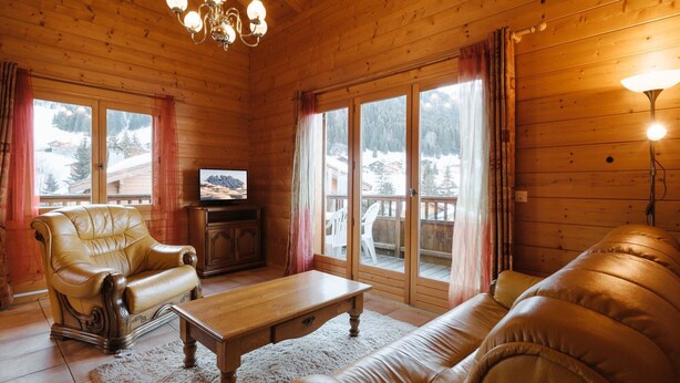 A cozy log cabin interior with a leather sofa, a wooden coffee table, a television on a cabinet, and large windows with curtains.