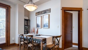 Un coin repas avec une table en bois, quatre chaises et un lustre. Il y a une porte sur la droite et une fenêtre avec des stores sur la gauche.