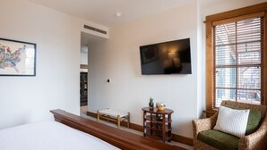 Une chambre d’hôtel avec un lit, un téléviseur fixé au mur, un fauteuil en osier avec un coussin, une petite table avec une plante et une carte encadrée accrochée au mur.