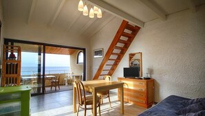 A room with a dining table, chairs, a TV on a wooden cabinet, a sofa, and a view of the sea through a glass door. - Nice 2-room Apt, 4 people, Lumio, AC, parking (Lumio)
