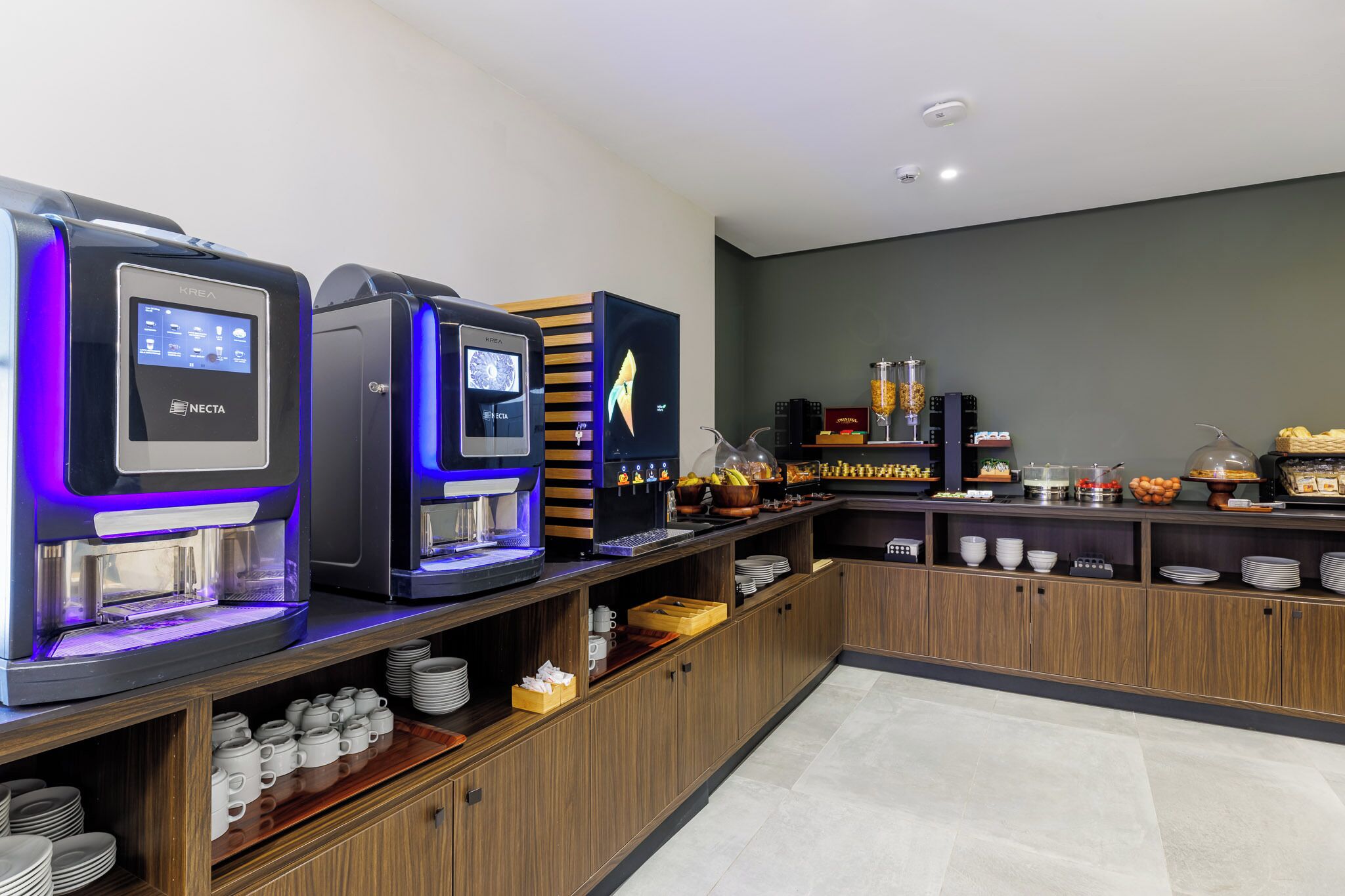 A self-service breakfast area with coffee machines, a fruit display, and various breakfast items.