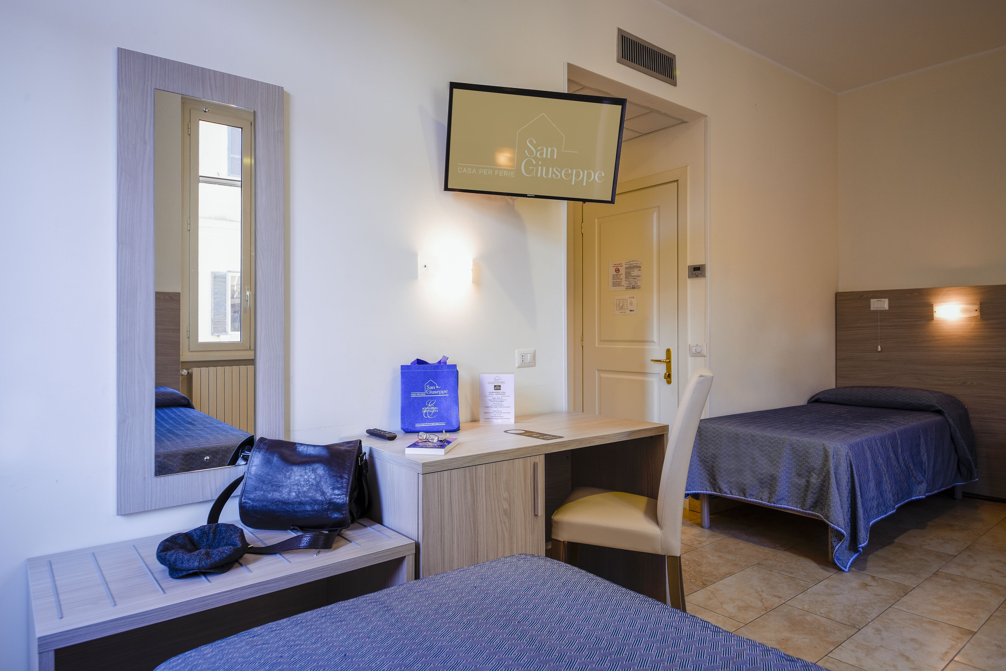 A hotel room with a bed, a desk, a chair, a mirror, and a television displaying "San Giuseppe".