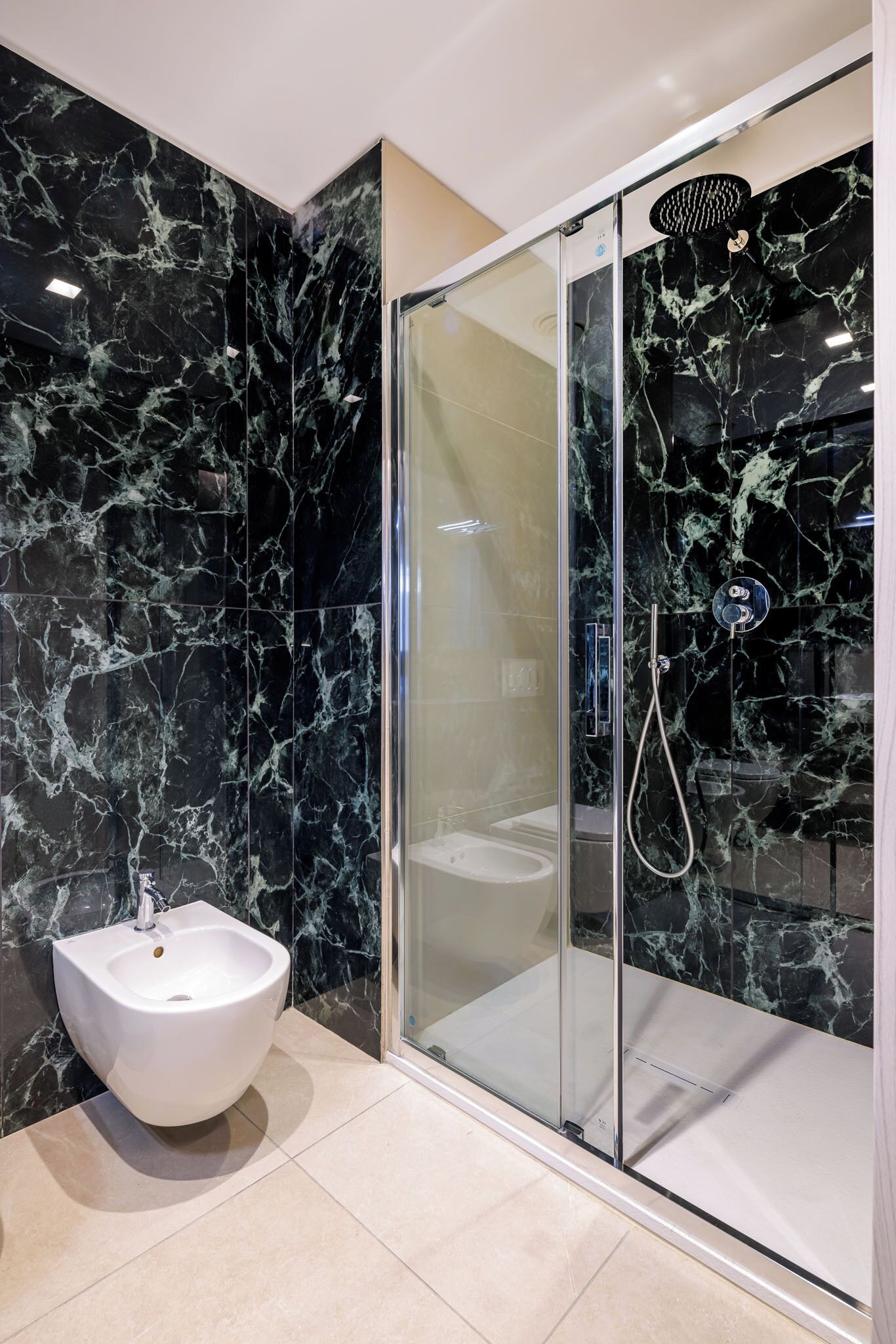 A modern bathroom with a glass-enclosed shower, a white pedestal sink, and marble walls.