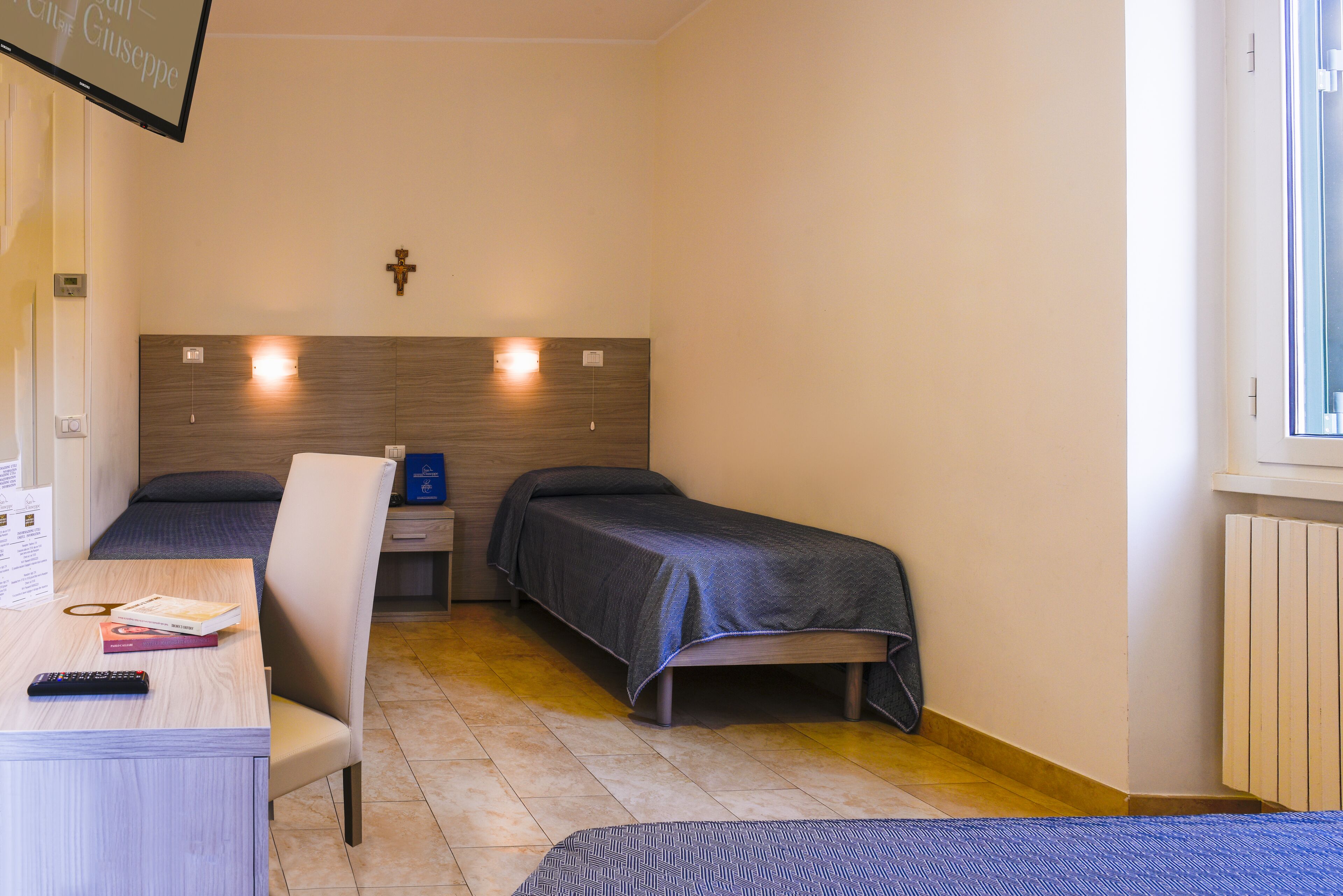 A room with two single beds, a desk with a chair, a TV mounted on the wall, and a window on the right.