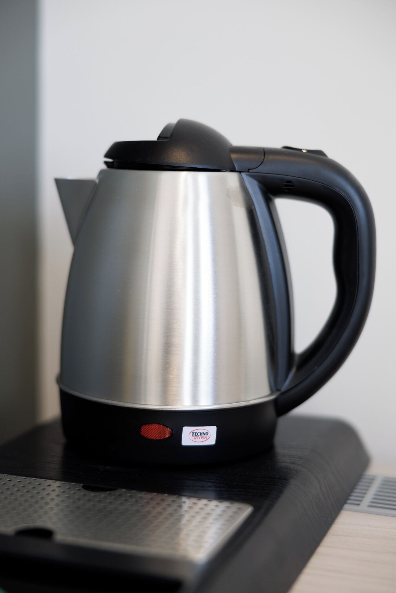 A stainless steel kettle with a black handle and a red button, placed on a black surface.