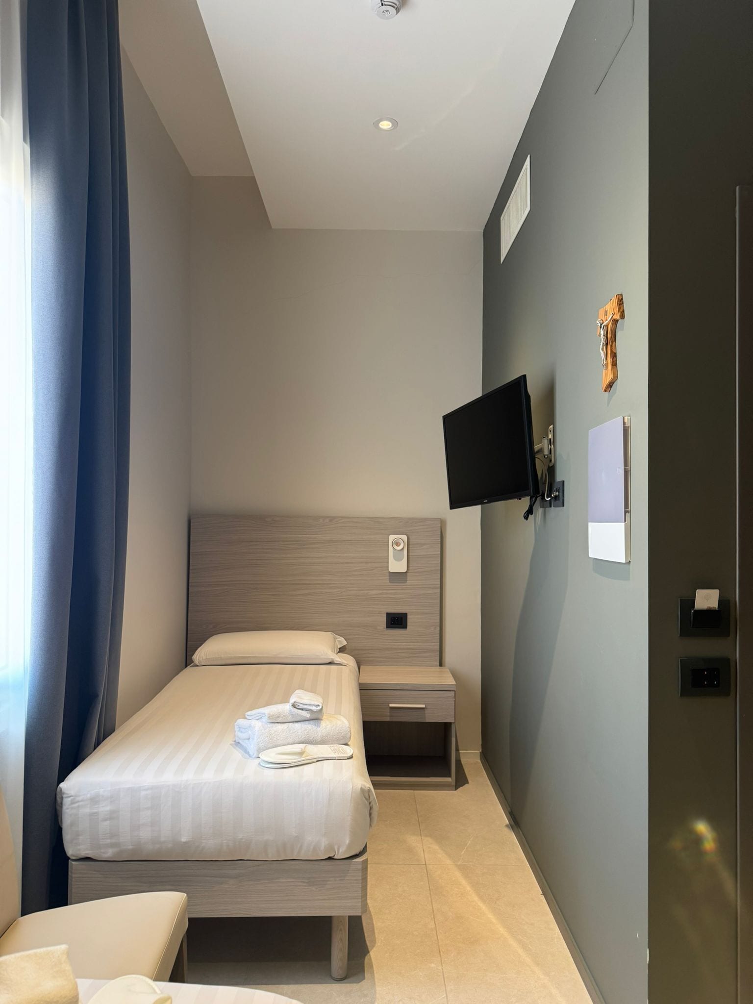 A hotel room with a bed, bedside table, TV, and a wall-mounted control panel.