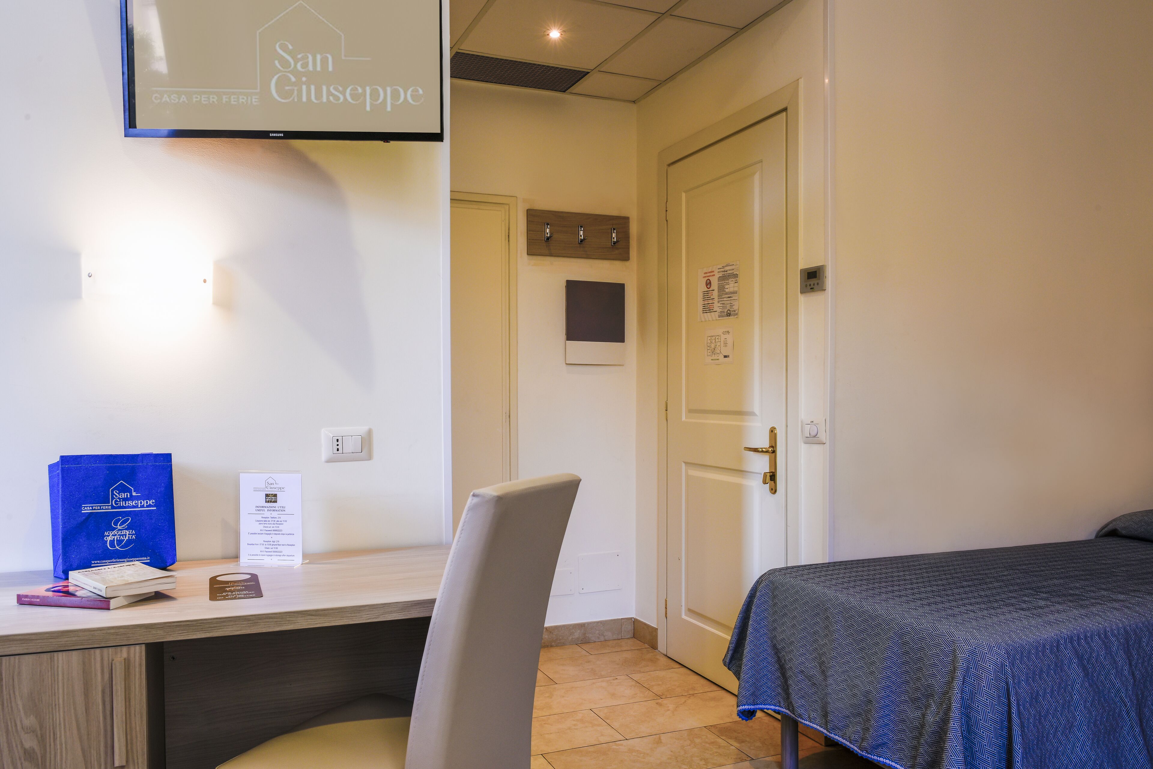 A hotel room with a bed, a desk with a chair, a TV mounted on the wall, and a door with a small window.