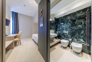 Habitación de hotel moderna con escritorio, silla, cama y baño con baldosas de mármol, bidés y lavamanos.