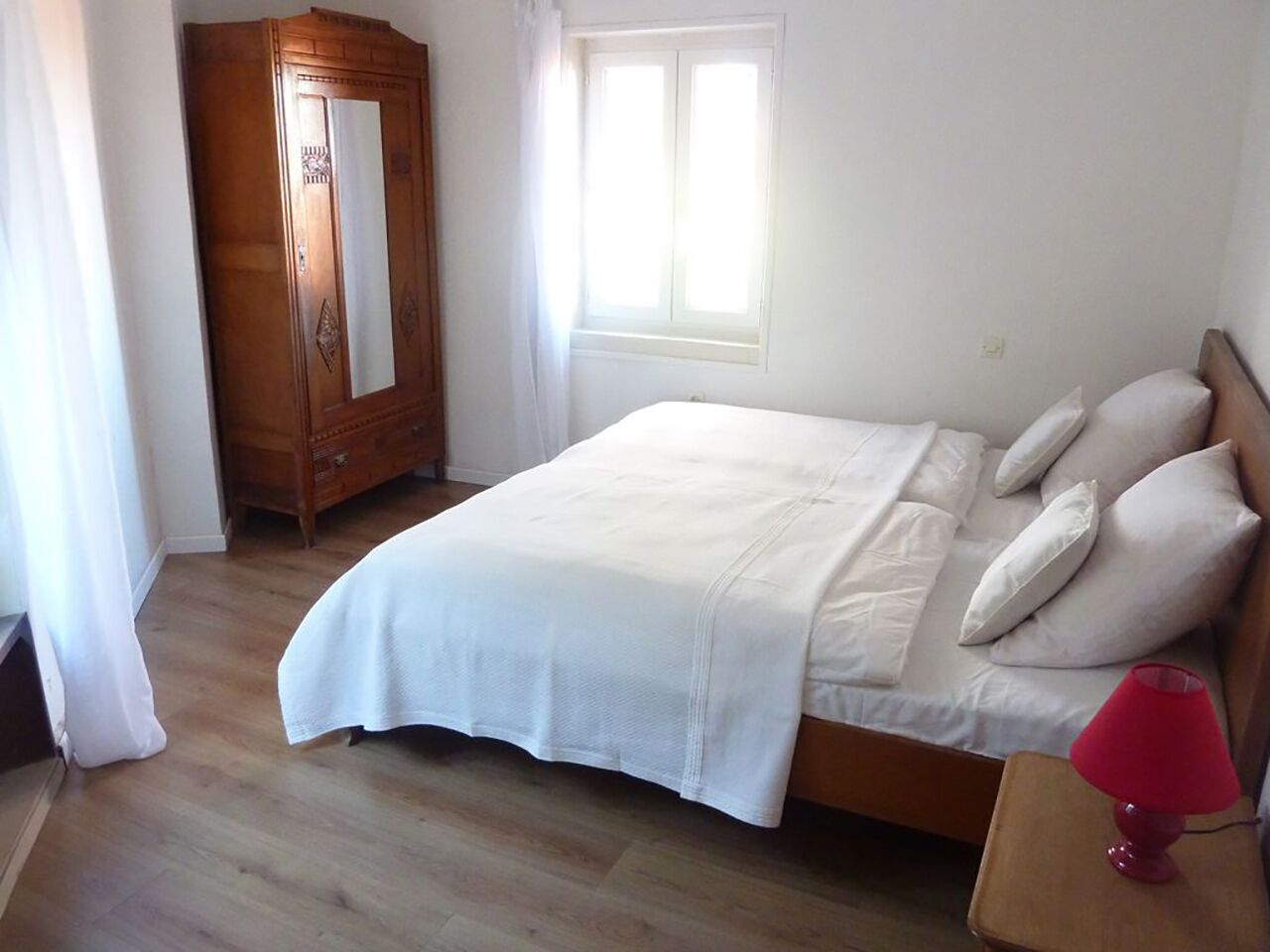 Quarto com cama, guarda-roupa de madeira, janela e abajur vermelho em uma mesinha de cabeceira.