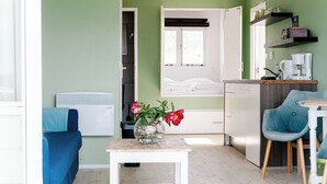 Ein gemütlicher Raum mit einem Bett, einem kleinen Tisch mit Blumen, einem blauen Stuhl, einer Küchenzeile und einem Fenster mit Jalousien.