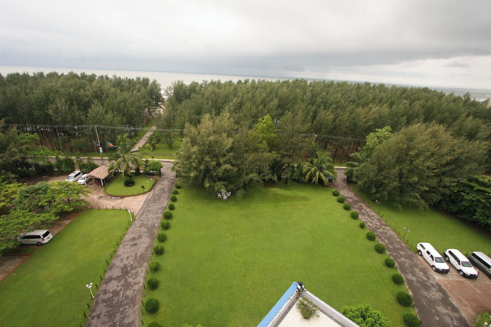 An aerial view of a property with a large green lawn, a parking area with several cars, and a dense forest in the background.