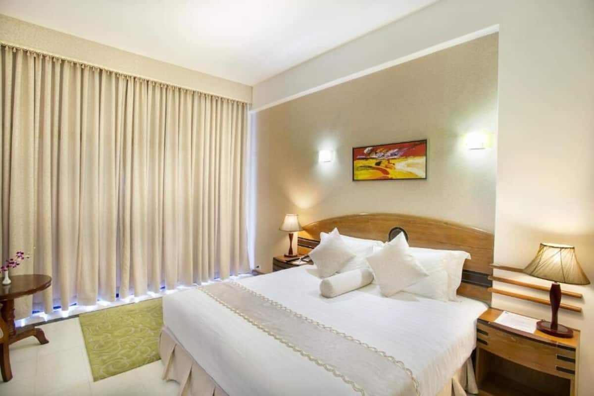 A hotel room with a large bed, a wooden headboard, a bedside table with a lamp, a small table with a vase of flowers, and a painting on the wall.