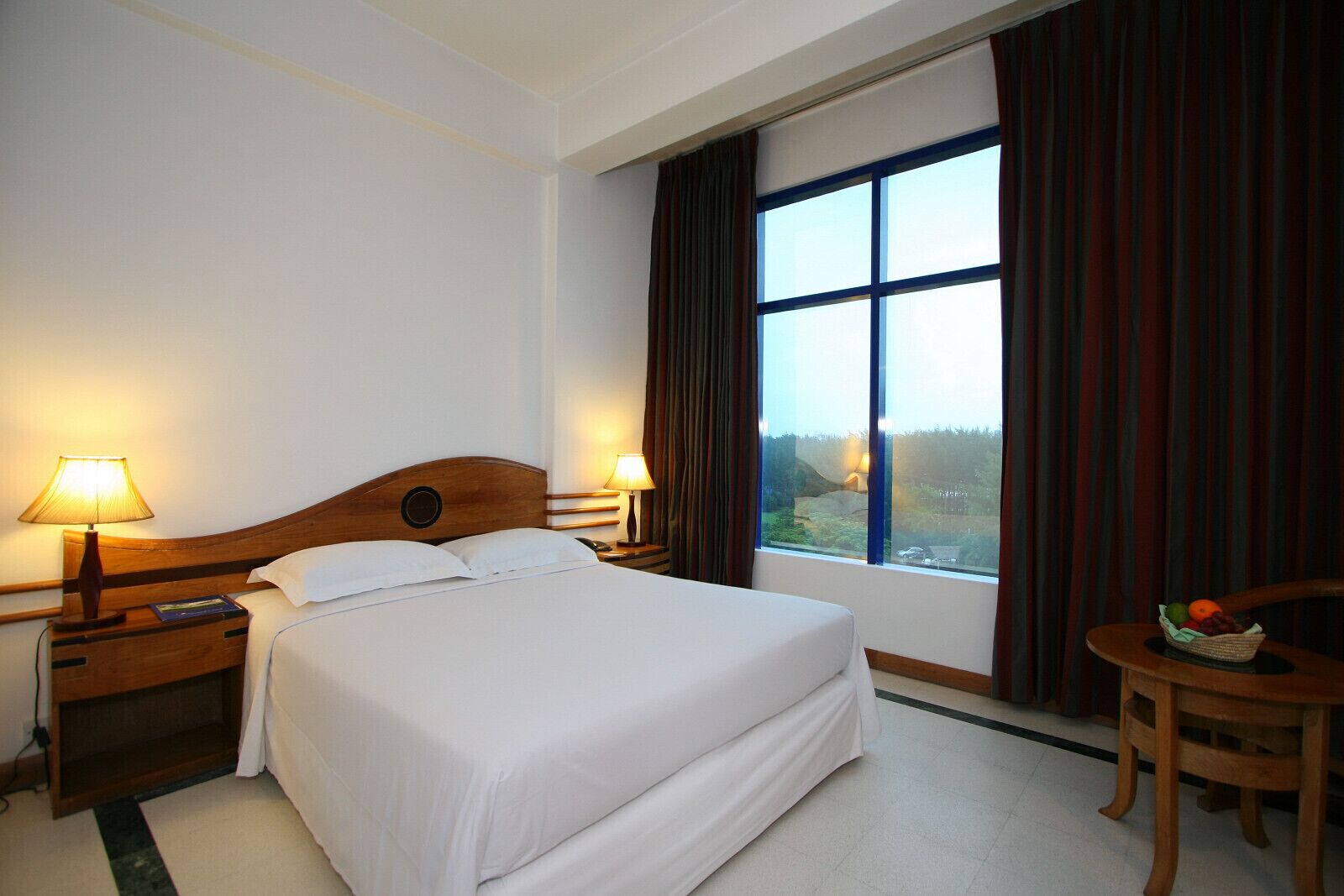 Una habitación de hotel con una cama grande, dos lámparas de noche, una mesita con fruta y una ventana con cortinas.