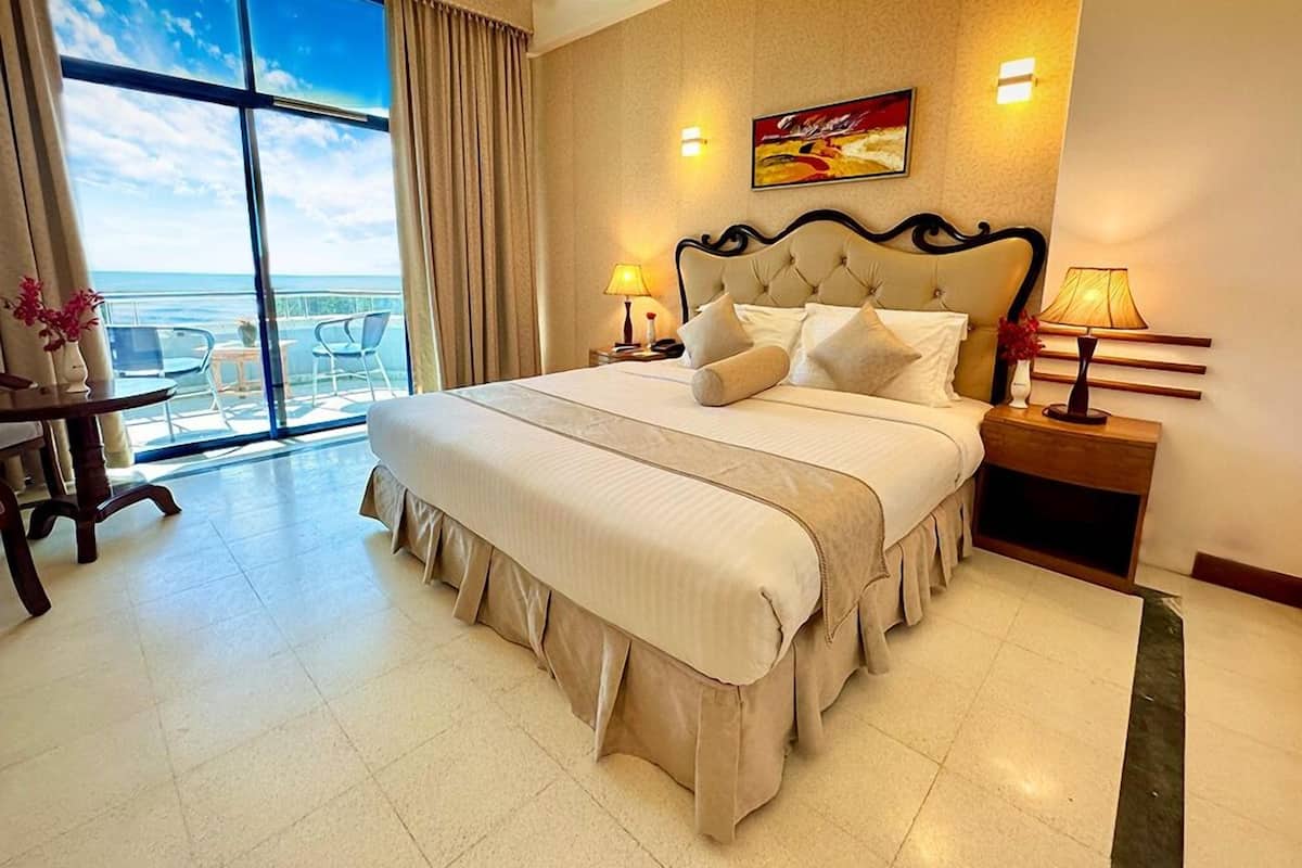 A spacious hotel room with a large bed, two bedside lamps, a view of the sea through large windows, and a balcony with chairs.