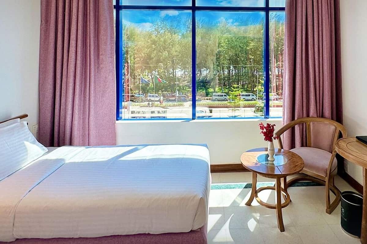 A hotel room with a bed, a chair, a small table, and a window with a view of a parking lot and trees.