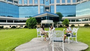 A modern building with a glass facade and a covered outdoor seating area with white chairs and a table. - Seagull Hotels Ltd. (Cox's Bazar)