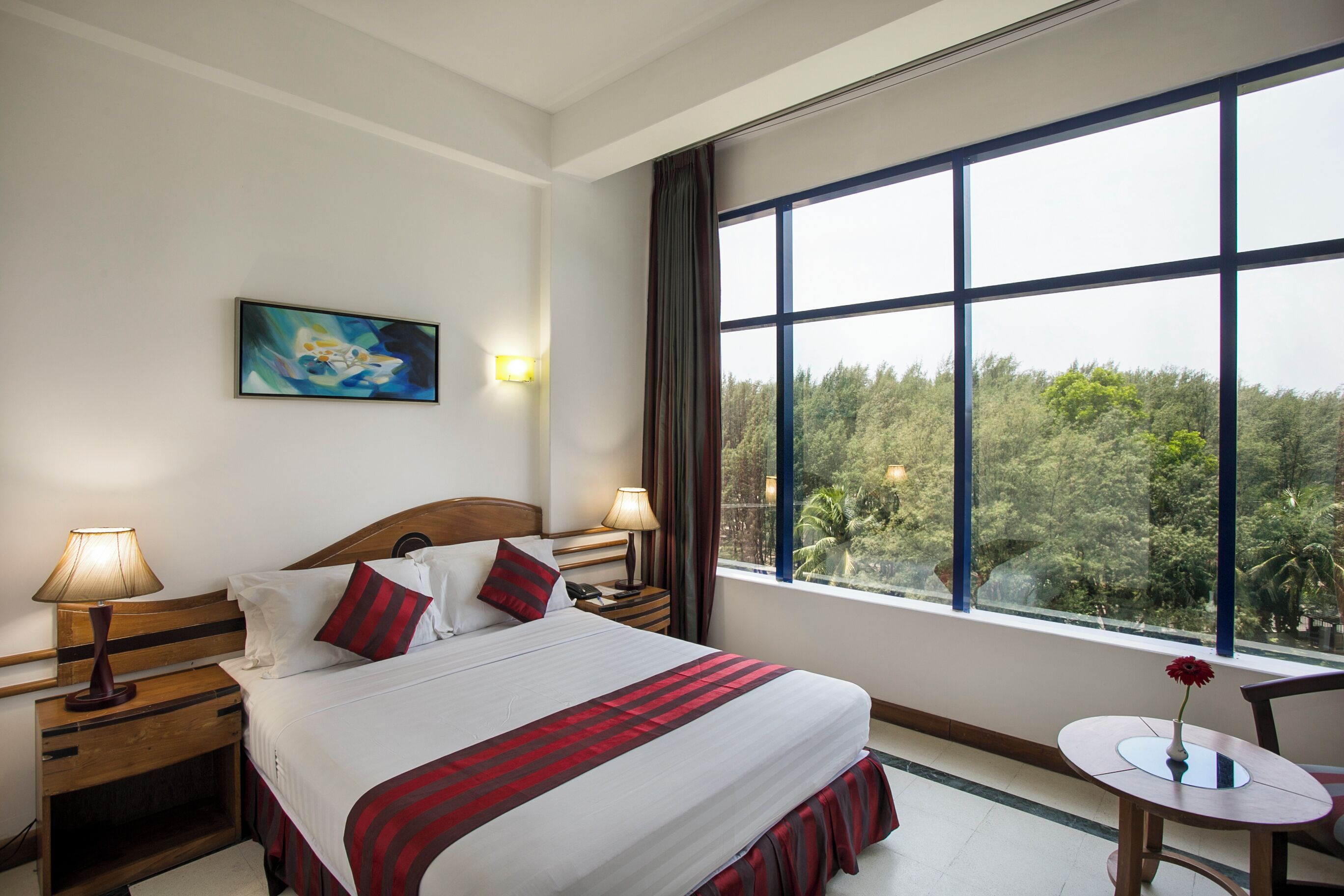 Habitació d'hotel amb un llit gran, dues taules de nit amb làmpades, una taula rodona petita amb un vas i una pintura a la paret, amb vistes a arbres a través de grans finestres.