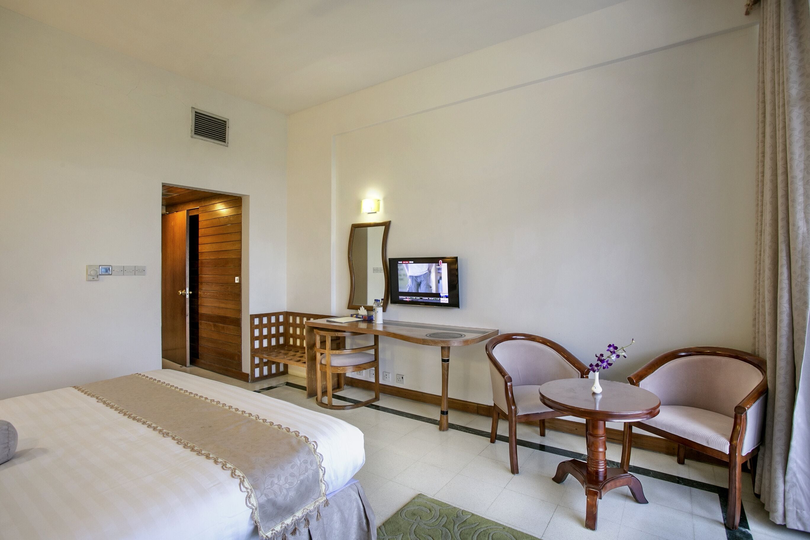 A hotel room with a bed, a desk with a chair, a television, and a small table with a vase.
