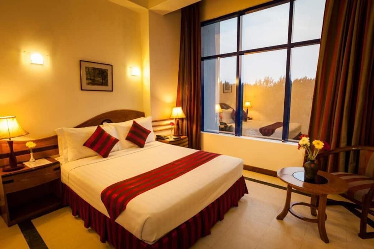 A hotel room with a large bed, two bedside tables with lamps, a small round table with a vase of flowers, and a window with curtains.