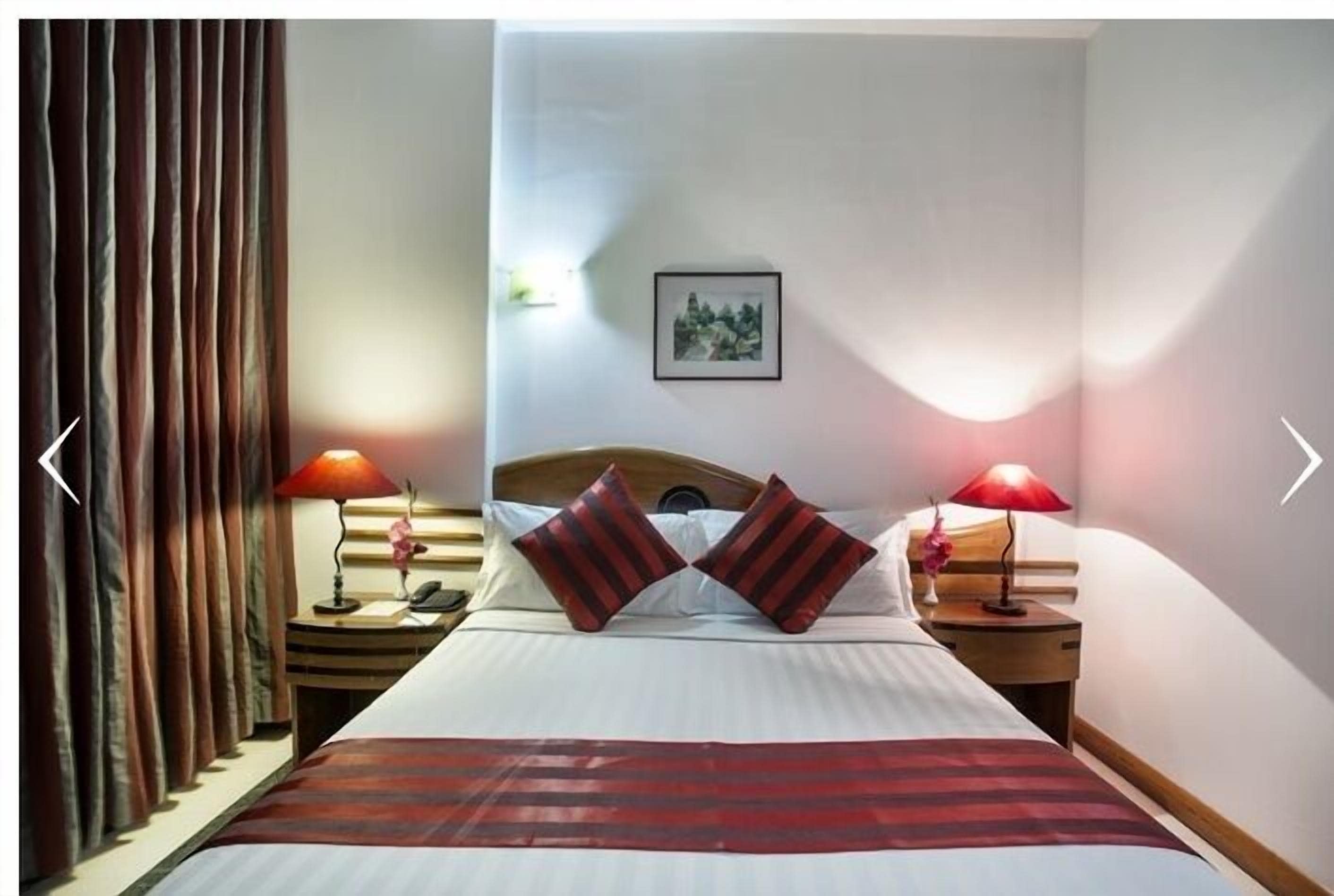A hotel room with a bed, two bedside lamps, a nightstand, a framed picture on the wall, and a window with curtains.