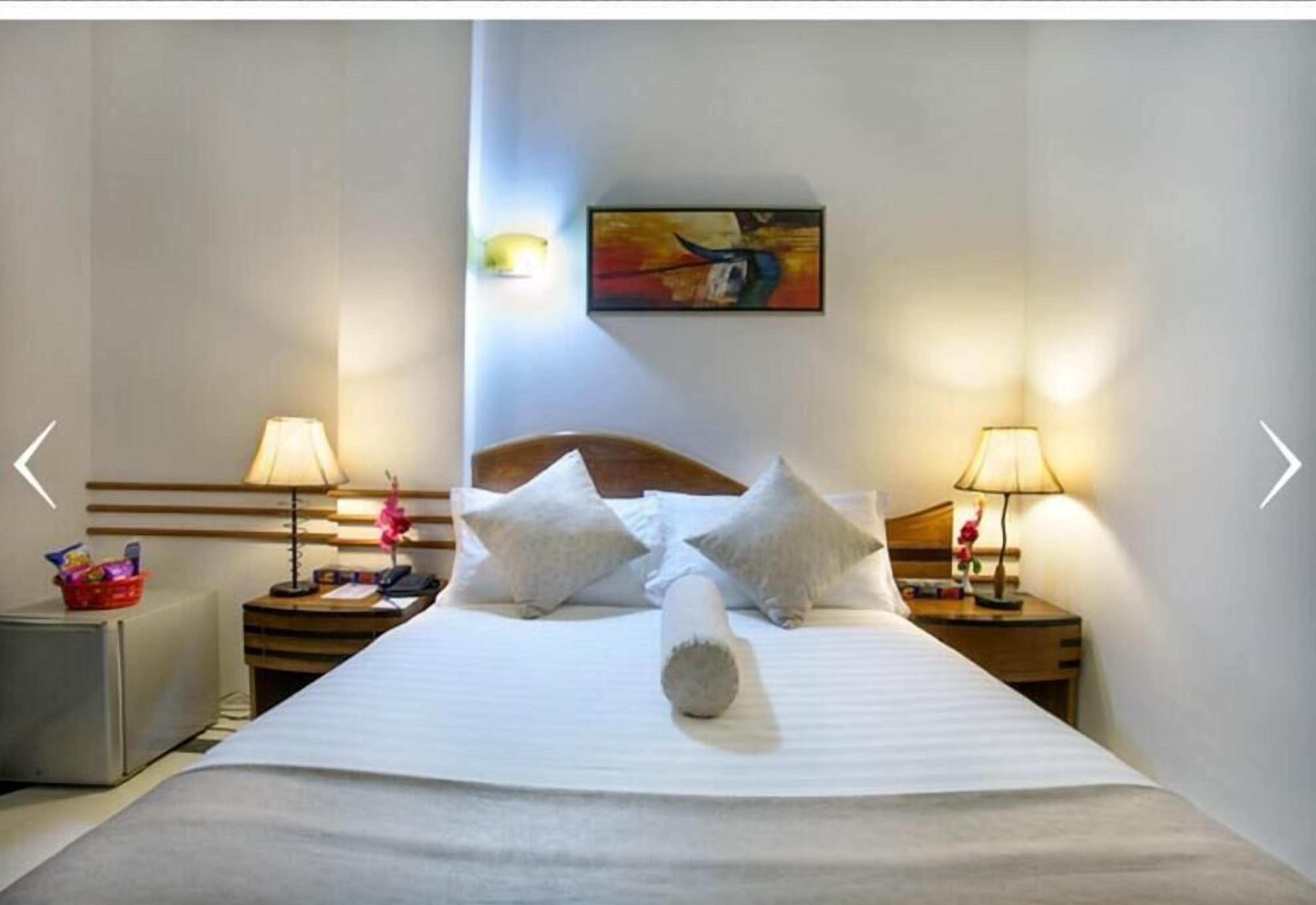 Hotellihuone, jossa on siististi pedattu sänky, kaksi yöpöytää valaisimineen, seinälle ripustettu taulu ja pieni jääkaappi vasemmalla puolella.