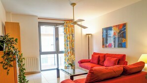 Ein modernes Wohnzimmer mit einem roten Sofa, einem Glastisch und einem Deckenventilator. Es gibt ein großes Fenster mit Vorhängen, eine Pflanze und ein Bild an der Wand.