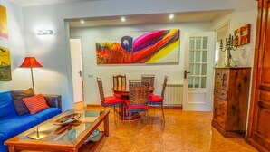 Un salon avec un canapé bleu, une table basse en verre, une table à manger avec des chaises, une grande peinture abstraite et un meuble en bois.