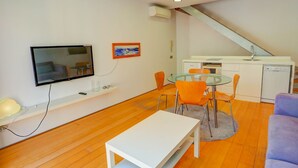 A modern living room with a flat-screen TV, a glass dining table with orange chairs, a white sofa, and a kitchen area with a sink and dishwasher. - Port Alegre Attic by Hello Homes Sitges (Sitges)