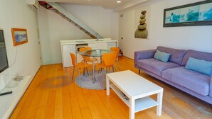 A modern living room with a purple sofa, a glass dining table with orange chairs, a white coffee table, and a staircase leading to a kitchen area. - Port Alegre Attic by Hello Homes Sitges (Sitges)
