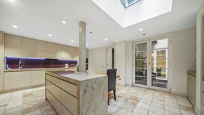 A modern kitchen with a central island, a dining area with a table and chairs, and a large sliding glass door leading to an outdoor space. - Magnificent House in Knightsbridge (London)