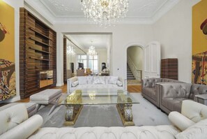 Un salon spacieux agrémenté d’un grand lustre, d’un canapé blanc, d’un canapé gris et d’une table basse en verre.