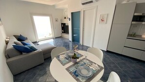 A modern living room with a grey sofa, a dining table with a decorative centerpiece, and a kitchen area with white cabinets and an air conditioning unit. - La Miravè holiday home located in Praiano, jewel of the Amalfi Coast. (Praiano)