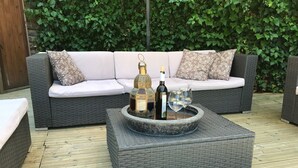 Un canapé en osier agrémenté de coussins à motifs, une table basse en pierre sur laquelle reposent une bouteille et des verres, et une terrasse en bois.
