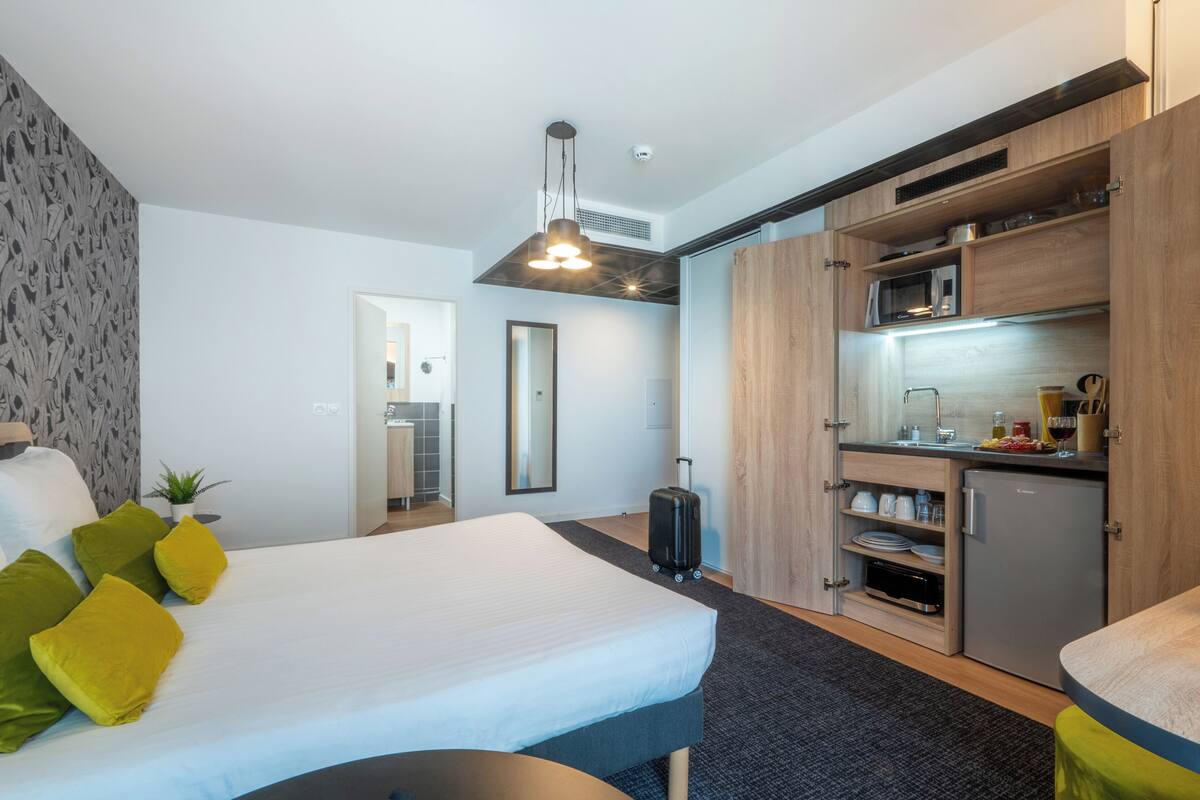 A hotel room with a bed, a small kitchenette, a suitcase, and a mirror.