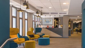 Un hall d’hôtel moderne avec des sièges jaunes et bleus, un comptoir d’accueil et de grandes fenêtres.
