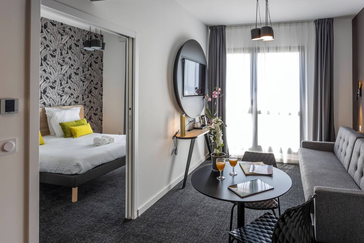 A hotel room with a bed, a sofa, a round table with drinks, a mirror, and a window with curtains.