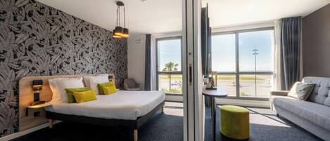 A hotel room with a bed, a sofa, a small table, and a view of the airport.