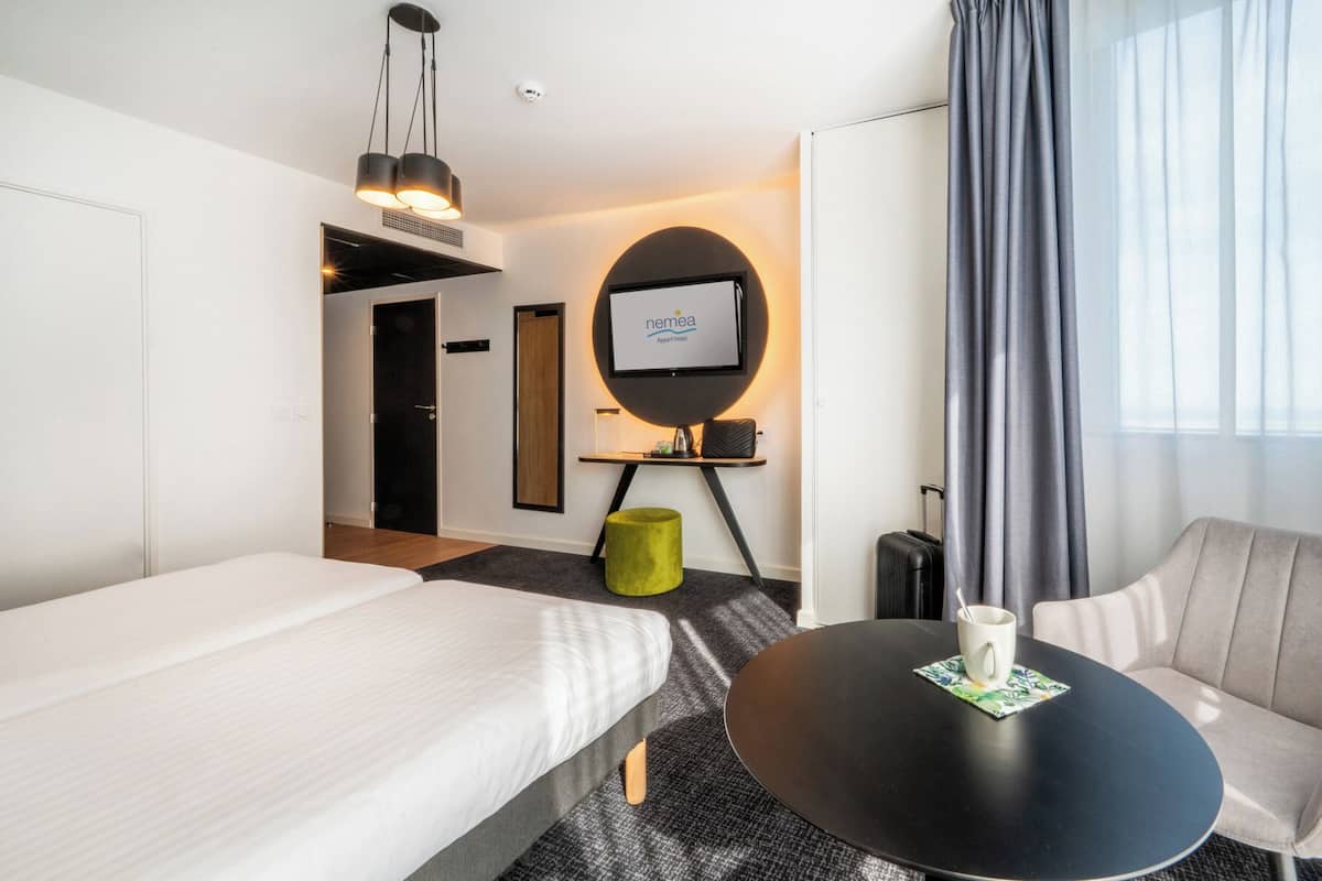 A hotel room with a bed, a round table with a cup, a green ottoman, a TV, and a window with curtains.