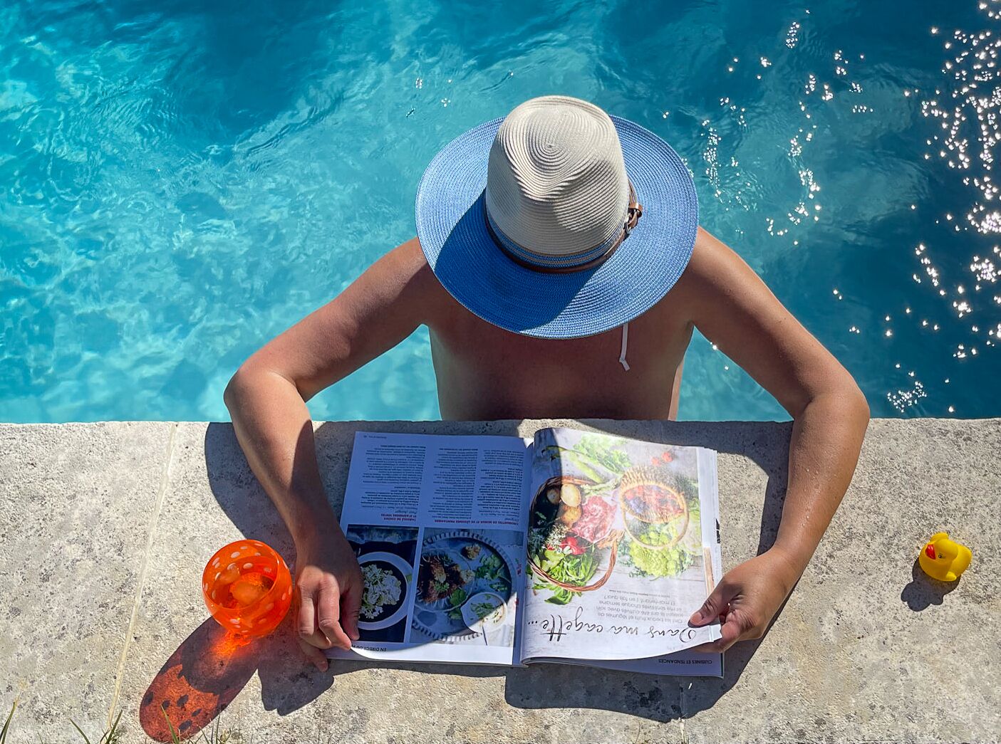 A person reading a magazine by a pool with a drink and a rubber duck.