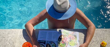 A person reading a magazine by a pool with a drink and a rubber duck.