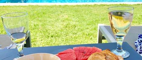 A table set outdoors with a pool view, featuring a variety of food including olives, cheese, and bread.