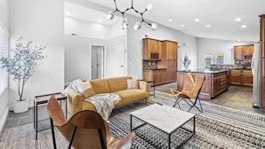 A spacious living room with a sofa, coffee table, and chairs. A kitchen with wooden cabinets and a central island is visible in the background. - The Scott With Heated Pool by Homeslice Stays (Scottsdale)
