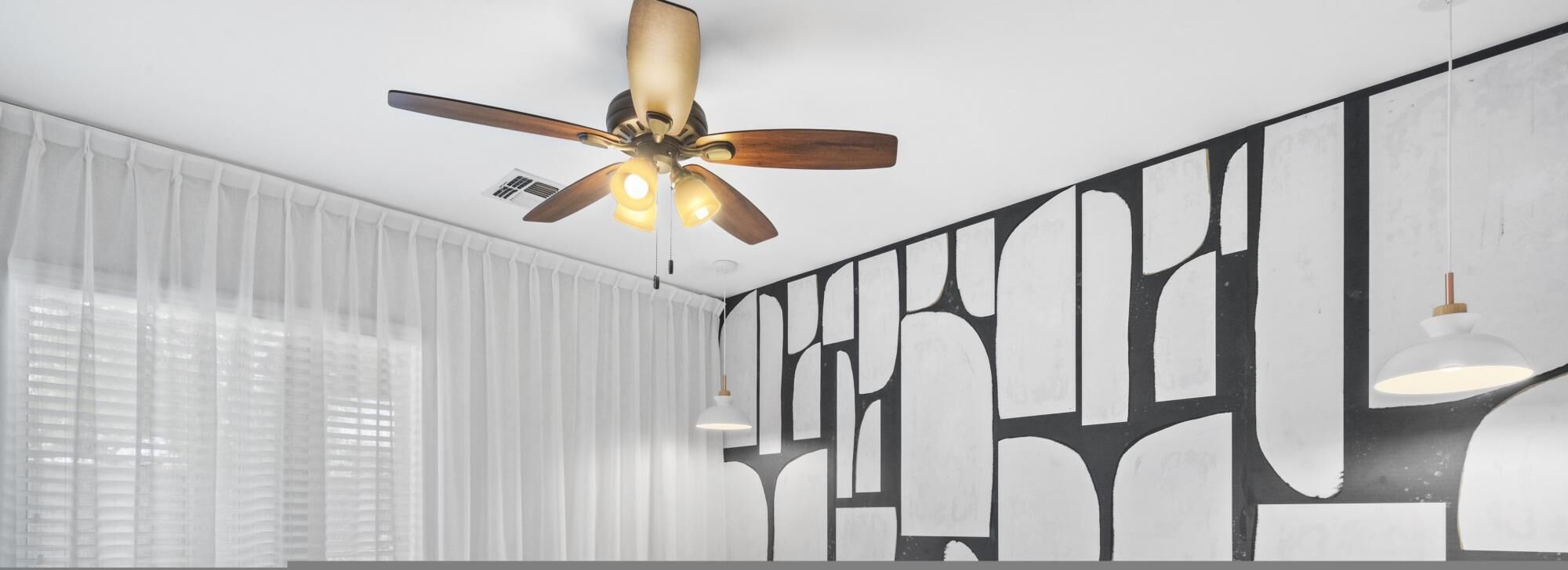 Una camera moderna con ventilatore a soffitto, ampie finestre con tende leggere e opere d'arte astratte alle pareti.