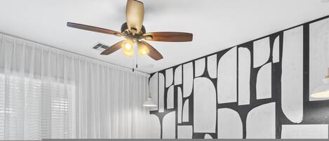 Una camera moderna con ventilatore a soffitto, ampie finestre con tende leggere e opere d'arte astratte alle pareti.