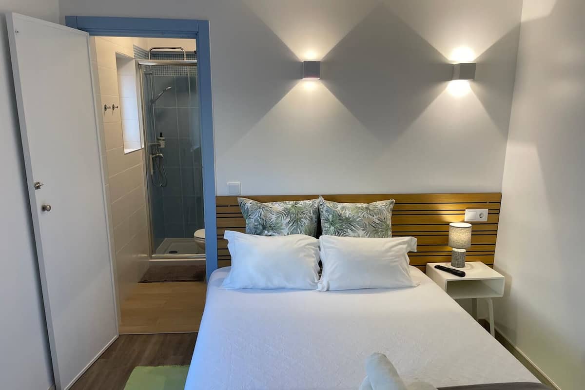 A bedroom with a bed, two pillows, a nightstand with a lamp, a shower area, and a door.