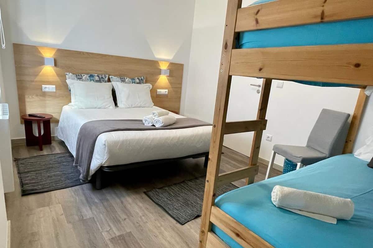 A hotel room with a bunk bed, a single bed, wooden headboard, bedside lamps, a small table, and a chair.