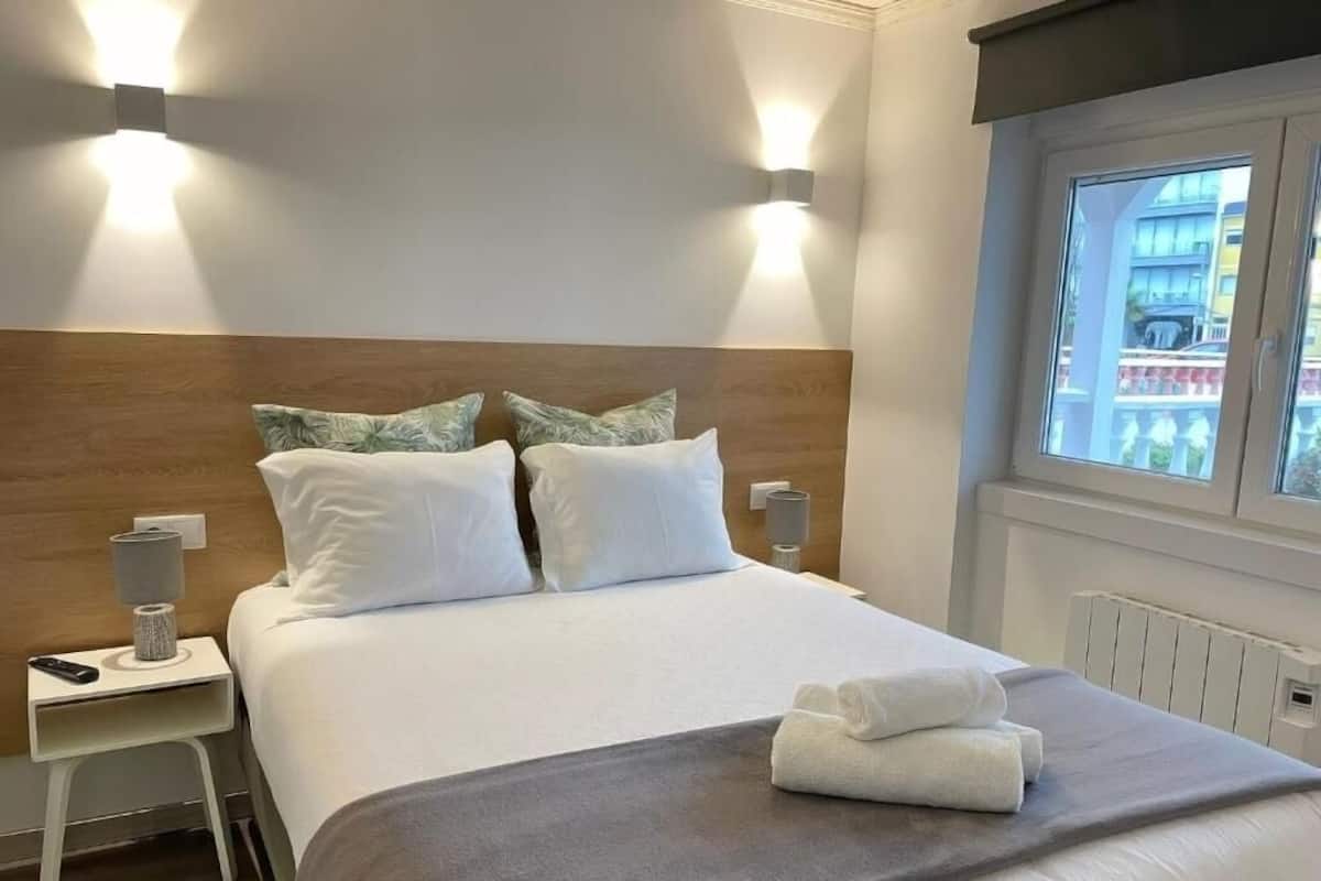A neatly made bed with white linens and a grey throw, bedside lamps, and a window with a view of buildings.