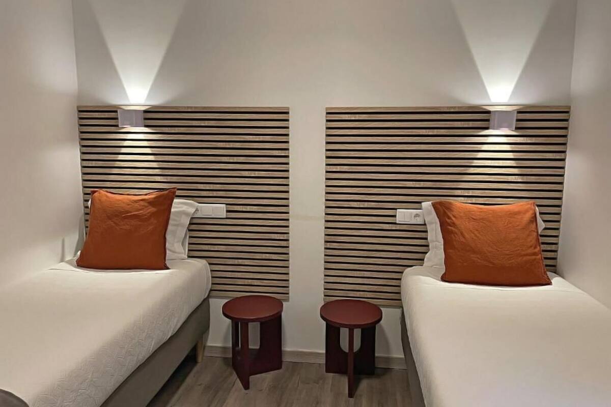 A hotel room with two beds, each with an orange pillow, wooden slat headboards, and small round tables.